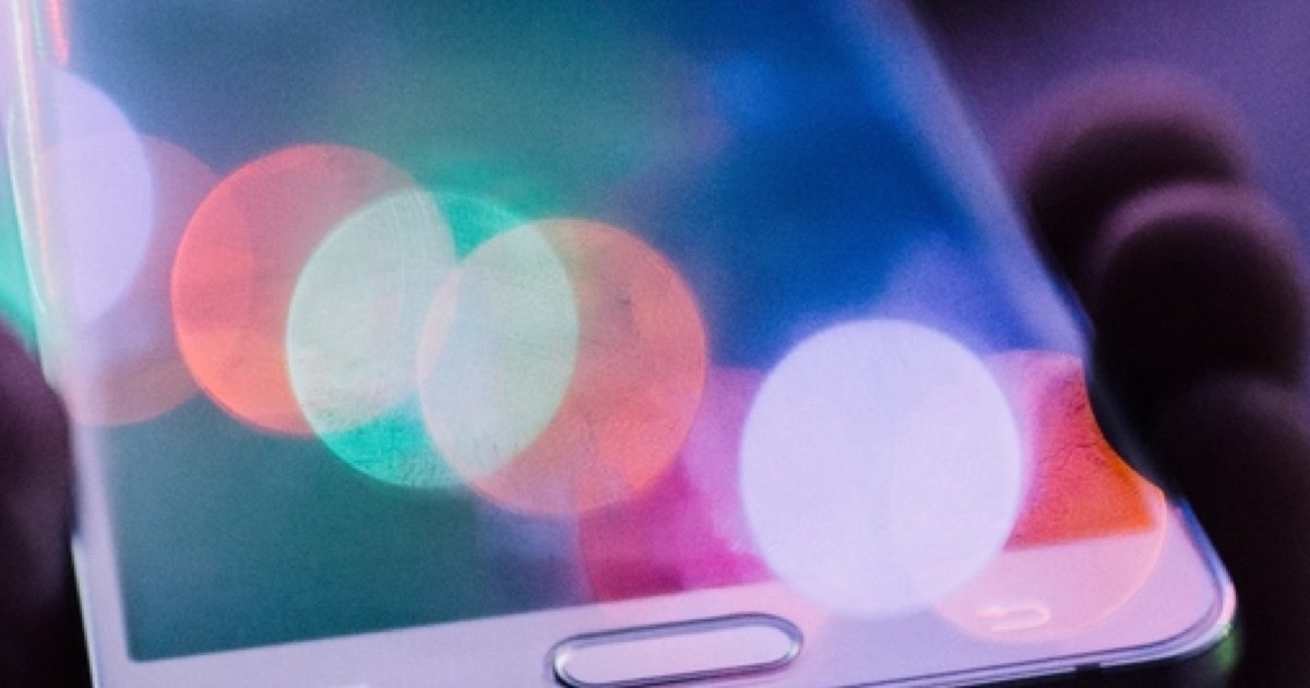 A close-up of a white smartphone held in a hand, its screen covered in colored bokeh lights