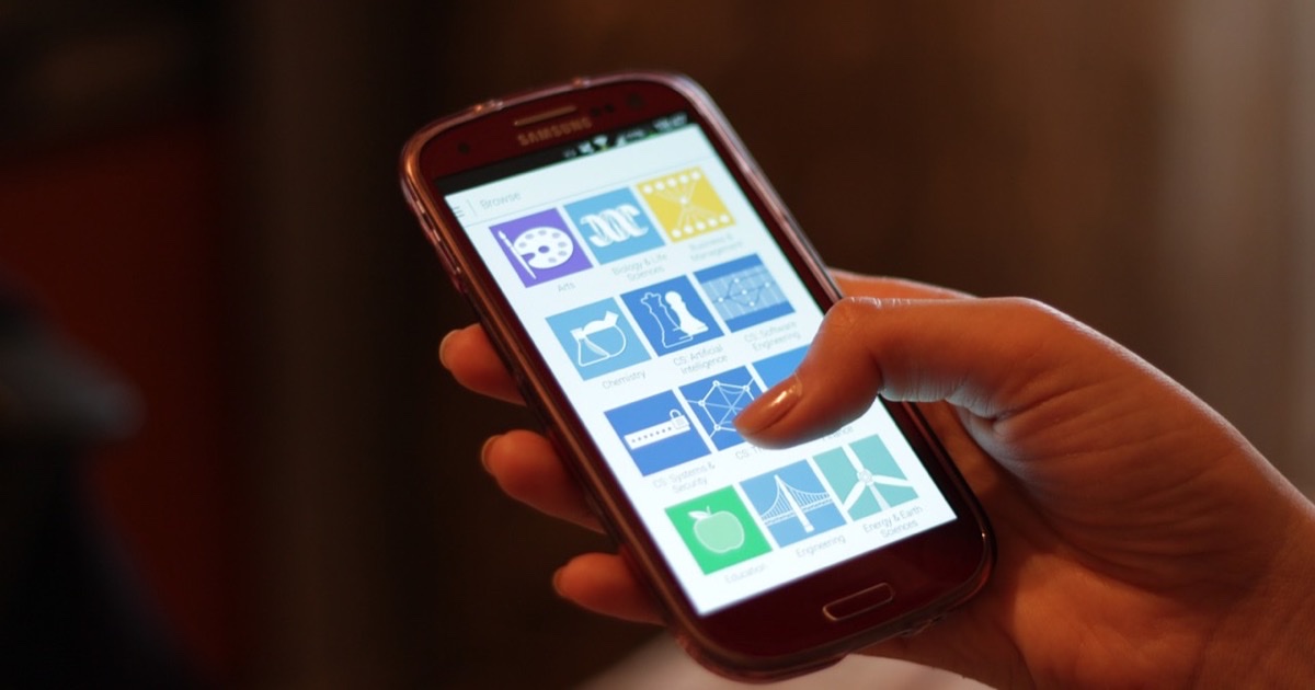 A hand holding a Samsung phone, a thumb resting on a grid of colored app tiles on the screen
