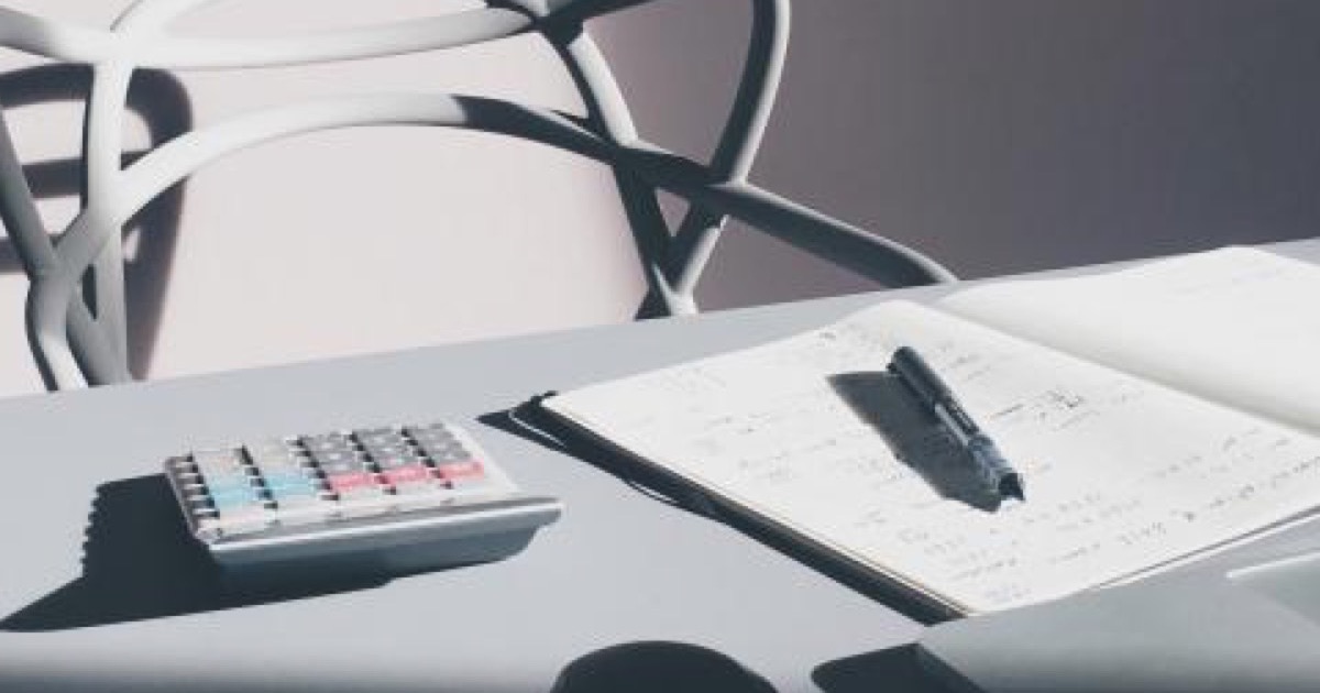 An open notebook, a pen and a calculator on a desk, a white chair behind