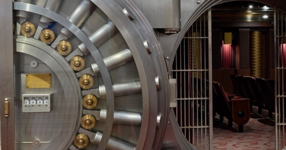 An open steel bank vault door with brass gears, a safe deposit room behind it