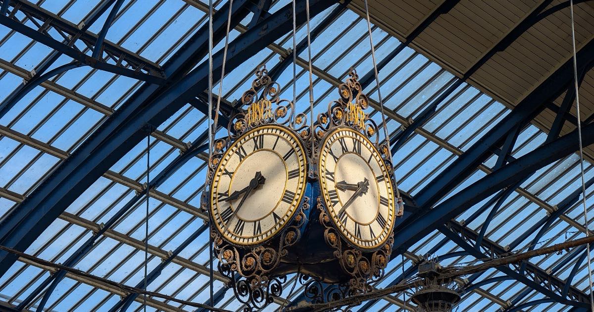 A twin-faced station clock in wrought iron hanging beneath a glass and steel roof