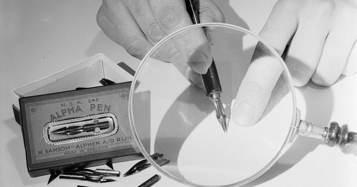 A pen nib held under a large magnifying lens, next to a box of spare nibs